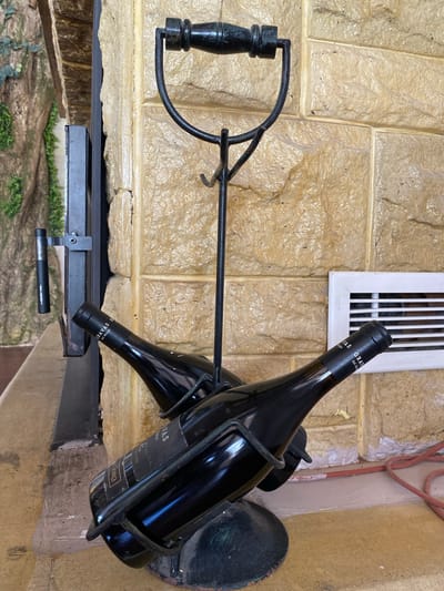A black metal wine rack holding two dark glass wine bottles stands on a textured, light-brown tiled surface against a similar tiled wall. The wine rack has a decorative handle at the top and a circular base. The wine bottles are positioned diagonally, with one bottle more prominently displayed in the foreground. The labels on the bottles are black with white text, partially obscured. To the left of the wine rack, a black metal structure with a handle is visible, possibly part of a door or gate. A white ventilation grate is mounted on the wall to the right of the wine rack. A red electrical cord lies on the floor in the lower right corner. The background to the left shows a textured, greenish-brown surface resembling artificial foliage or a painted mural. The scene appears to be outdoors or in a covered outdoor area, possibly a patio or entrance. The location is identified as Kyrenia, Northern Cyprus / Cyprus.