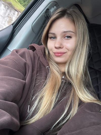 A young woman with long, blonde hair poses for a selfie in what appears to be the passenger seat of a car. She wears a brown hooded sweatshirt with a zippered front. The car's interior is visible, including a quilted headrest and a car door with a window. Outside the window, blurry greenery suggests the car is in motion. The location is not clearly identifiable as the city of Achadara, Abkhazia.