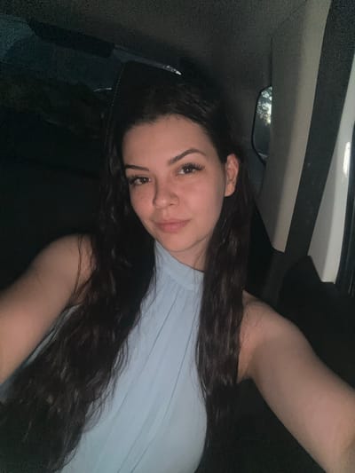 A young woman with long, dark hair and fair skin is pictured in what appears to be the interior of a vehicle at night. She is wearing a light blue top with a high neckline. The woman is looking towards the camera, and a subtle smile can be seen on her face. Her right arm is visible, resting slightly forward. The background is dark and indistinct, suggesting low-light conditions, with some light reflecting off the vehicle's interior. There are no visible landmarks or contextual clues to place the image in Zlati Voyvoda, Bulgaria.