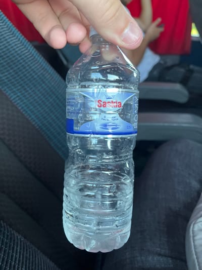 A person's hand holds a plastic bottle of water. The bottle is clear and contains a clear liquid with condensation on the outside. A blue and white label on the bottle displays the brand name "Saskia" in red lettering. The label also shows text that reads "Bronwater Eau de source" and "0,5 l". The background is out of focus but appears to be inside a vehicle, with dark seats and a red-colored object. The scene suggests a person is traveling or spending time in a car.