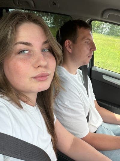 A young woman with long brown hair and blue eyes, and a man with short brown hair and brown eyes are sitting in the front seats of a car. The woman is in the foreground, looking at the camera, with her seatbelt across her chest. The man is in the background, looking out the window. Both are wearing white t-shirts. The car is moving, as indicated by the blurred greenery outside the window, suggesting a rural or suburban setting. The image does not provide any specific details about the city of Alnashi, Russia.