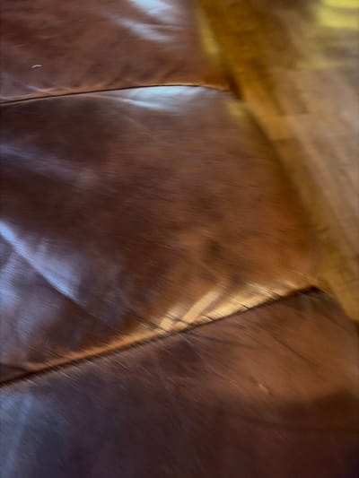 The image shows a close-up, abstract view of a brown leather surface, likely part of a piece of furniture such as a sofa or chair. The texture of the leather is visible, with slight creasing and variations in sheen due to lighting. There are seams running across the surface, dividing it into sections. In the upper left portion of the image, there appears to be a wooden surface, possibly a floor or table, with a warm, golden-brown hue. The lighting suggests an indoor environment, with light casting shadows and highlights on the leather. No people, animals, or specific activities are depicted. The scene is static and focuses solely on the material and textural qualities of the leather and wood. The colors are predominantly warm browns and golds. There is no visible text in the image. The location context of Stīveri, Latvia, does not provide specific visual cues within the image itself, but implies an everyday domestic or public setting.