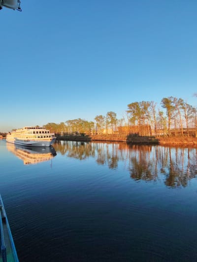 The image shows a river landscape in Myshkin, Russia. A large, white cruise ship is seen on the left side of the river, with its reflection visible in the water. Along the opposite bank of the river, a line of trees with sparse green foliage is reflected in the water. The sky is clear and blue. The water appears calm, with a dark to light blue gradient. A portion of another ship can be seen in the bottom-left corner of the picture. The scene suggests early morning or late afternoon lighting.