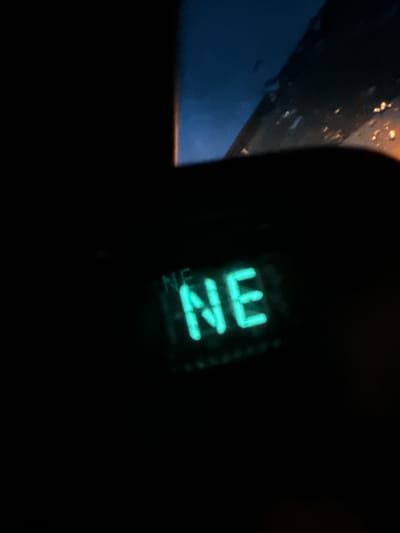 The image shows a close-up, dimly lit view of a digital display that reads "NE" in a bright green, neon-like font. The display appears to be part of a car's interior, possibly a compass or a navigation system, as suggested by the faint outlines of what could be a dashboard and a windshield with water droplets and distant lights visible through it. The overall scene is dark, with the exception of the glowing "NE" and some blurred orange lights in the background. There are no people or other discernible objects in the frame. The image was taken from inside a vehicle, likely at night or in very low light conditions. The specific location of Stony Plain, Canada cannot be determined from the visual information presented in the image.