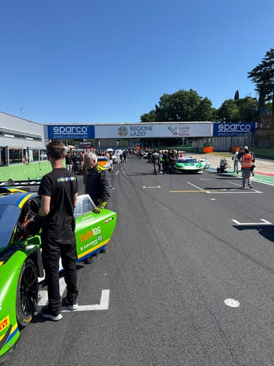 The image shows a scene at the Vallelunga Circuit, located in Campagnano di Roma, Italy. Several race cars are lined up on the track, likely preparing for a race or qualifying session. Two men are interacting with one of the cars in the foreground, appearing to be making adjustments or checks. The car is green, blue, and yellow with visible sponsor logos like "twis loc," "E Leclerc CAEN," "astron," "PERTAMINA," and "ELLI."  Banners for "sparco" and "REGIONE LAZIO" are visible on the side of the track. Other team members and spectators are gathered along the track barrier. The track surface is asphalt, marked with starting grid numbers. The weather appears sunny.