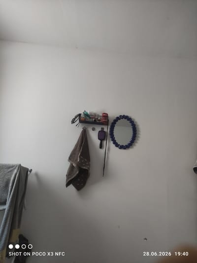 The image displays a section of a plain, off-white wall, likely in a bathroom or dressing area. On the left side, two grey towels are hung on a rod. To the right, a small shelf holds various items, including what appear to be toiletries and a pair of sunglasses. Below the shelf, a brown towel with a patterned edge hangs from a hook. Further to the right, a black hairbrush with a purple bristle head is suspended by its handle, and a round mirror with a dark blue, coiled frame is mounted on the wall. The bottom left corner of the image features a watermark indicating "SHOT ON POCO X3 NFC," and the bottom right corner displays a date and time stamp: "28.06.2026 19:40." The image is devoid of any people or discernible activity. The overall impression is one of a tidy, functional space.
