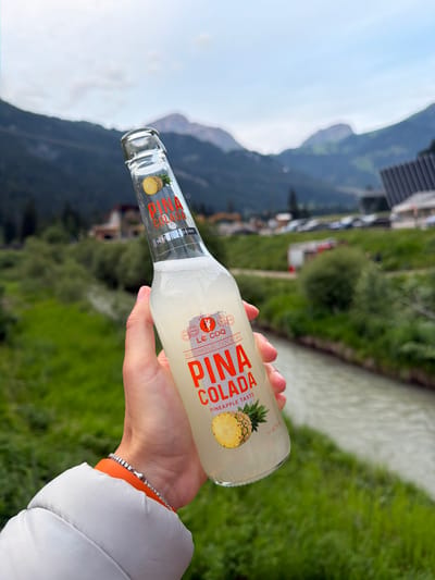 A hand holds a bottle of "Pina Colada" alcoholic cocktail against a backdrop of a scenic mountain landscape. The bottle's label prominently features the drink's name in orange text, along with an image of a pineapple and the text "Le Coq Alcoholic Cocktail" and "Pineapple Taste". The background showcases lush green grassy hills, a flowing river, and distant mountains with a cloudy sky overhead. Several buildings and parked cars are visible in the mid-ground, suggesting a populated area within the mountainous region. The person is wearing a gray puffy jacket and an orange accessory on their wrist, possibly a bracelet or watchband. The overall scene captures a relaxed moment in a beautiful, natural setting within the town of Campitello di Fassa, Italy, known for its picturesque mountain views and outdoor activities.