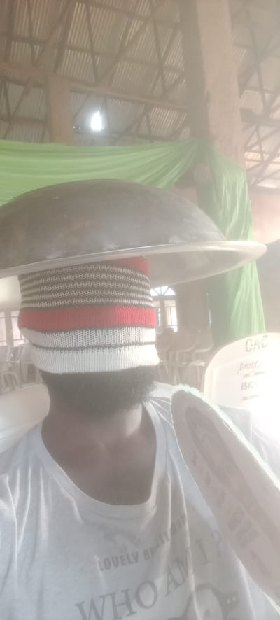 A man with a beard is wearing a knit cap covering his eyes, with a large, dark pan resting on top of his head. He is wearing a white t-shirt with the words "LOVELY GENTLEMAN" and "WHO AM I?" printed on it. The background shows what appears to be an interior space, possibly a church or event hall, with rows of white chairs, green draped fabric, and a corrugated metal roof. The environment is dusty or hazy. A chair in the background reads, "CAC AMOGU BENDE". The scene is taking place in Bende, Nigeria.