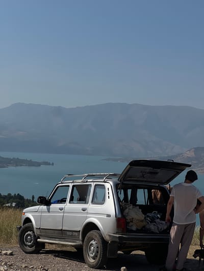A silver Niva vehicle is parked on a gravelly embankment overlooking a large, turquoise body of water, likely a reservoir. The vehicle's rear hatch is open, revealing the interior filled with various items, including bags and folded clothing. A young man, wearing a white mesh t-shirt and striped pants, stands by the open hatch, facing away from the camera and looking towards the water. In the background, hazy mountains rise majestically, with a lighter blue sky above. The environment appears dry and arid, with sparse vegetation on the embankment. A small dog is visible near the man's feet. The scene suggests a stop during a road trip or an outdoor excursion. The location is described as the city of Yangikurgan, Uzbekistan.