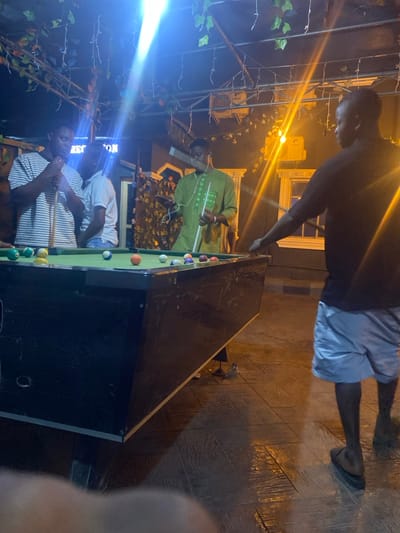 The image depicts four adult males engaged in or observing a game of pool at night within an outdoor or semi-outdoor recreational area in Igbessa, Nigeria, possibly a lounge or bar given a visible "RECEPTION" sign. A standard-sized pool table with a green felt playing surface occupies the central foreground, featuring numerous colored pool balls.
On the left, one male in a white and grey striped t-shirt holds a pool cue, appearing to observe the table. Partially obscured behind him is another male in a light-colored shirt, also looking towards the table. A third male, positioned center-right, wears a green long-sleeved garment; he holds a pool cue in his right hand and a mobile device with attached earbuds in his left hand. On the far right, a fourth male, viewed from the back, wears a dark t-shirt, light-colored shorts, and dark sandals; his right hand is extended towards the pool table.
The environment is illuminated by multiple artificial light sources, including a prominent blue-white overhead beam and warmer ambient lights in the background. An overhead trellis structure is adorned with artificial green foliage and small decorative lights. The ground is paved with patterned tiles, exhibiting reflections from the lighting. Architectural elements resembling pillars and window frames are discernible in the background.
