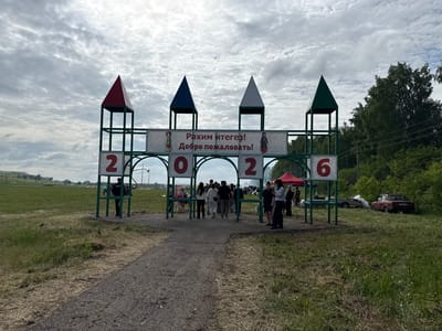 An outdoor event entrance structure is set up on a grassy field under a cloudy sky. The structure consists of four green archways, each topped with a conical roof in red, blue, white, and green. Large numbers are displayed on the archways: '2', '0', '2', and '6'. A banner across the top of the structure reads "Рәхим итегез! Добро пожаловать!" (Welcome! in Tatar and Russian), with images of two traditional dolls on either side. A group of people are walking through the archways, suggesting they are entering an event. In the background, a red car is parked to the right, and beyond that, a dense line of trees borders the field. The overall scene suggests a community gathering or festival in Старое Субханкулово, Russia.