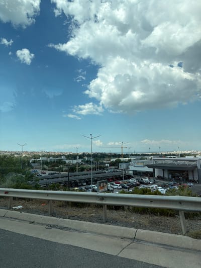 This image captures a panoramic view of a suburban area in Efkarpia Municipal Unit, Greece, dominated by a large retail establishment with an extensive parking lot filled with vehicles. The scene is set against a backdrop of a bright blue sky dotted with fluffy white clouds. In the foreground, a concrete barrier and a guardrail line a road, suggesting a vantage point from an elevated position, possibly a bridge or overpass.

The retail building, likely a supermarket or shopping center, is modern in design, featuring a large, flat roof structure and prominent signage that reads "KOUS-KOUS." Cars of various colors and models are densely packed in the outdoor parking area, which appears to be partially covered by a canopy system, possibly for solar panels or shade. The surrounding landscape is a mix of sparse vegetation and scattered buildings, indicating urban development.

Several streetlights with multiple fixtures are visible throughout the scene, along with what appears to be construction cranes in the distance, hinting at ongoing development in the area. The overall impression is one of a typical commercial hub within a Greek suburban setting, characterized by its functional architecture and extensive infrastructure for consumer access. No individuals are clearly distinguishable in the frame, emphasizing the focus on the built environment and infrastructure.