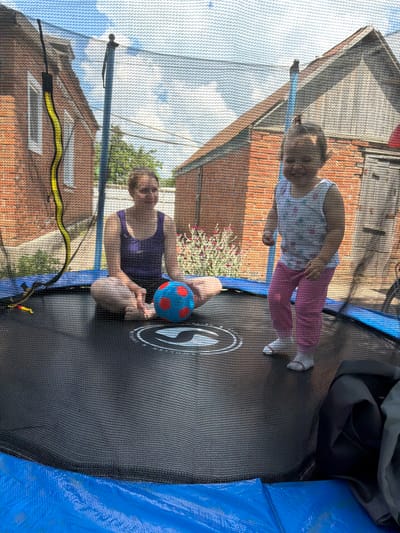 In Komsomolskiy, Russia, a woman and a toddler are on a trampoline. The woman, seated cross-legged, wears a dark purple tank top and dark shorts. A blue and red ball rests between her legs. The toddler, standing, is dressed in a white floral tank top and pink pants, with white socks. They are in a backyard, with brick houses visible in the background. The sky is blue with scattered clouds.