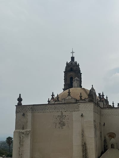 A photograph captures a grand, light-colored historical building, likely a church or monastery, set against an overcast sky in Tepotzotlán, Mexico. The main structure, constructed of aged stone or stucco, features intricate architectural details, including decorative friezes and finials along its cornices.
A prominent domed section is visible, with ornate carvings at its base. Behind this, a tall, elaborately decorated bell tower rises, topped with a cross. The tower displays multiple tiers and arched openings, characteristic of colonial Spanish architecture. The sky is a uniform, soft grey, indicating an overcast day.
Details on the building's facade include what appear to be drainpipes or similar fixtures and a small, circular window. In the lower right corner, a palm tree and distant foliage suggest a broader landscape setting beyond the immediate structure. No people are visible, and no specific activity is taking place; the image is a static architectural study. There is no visible text.
