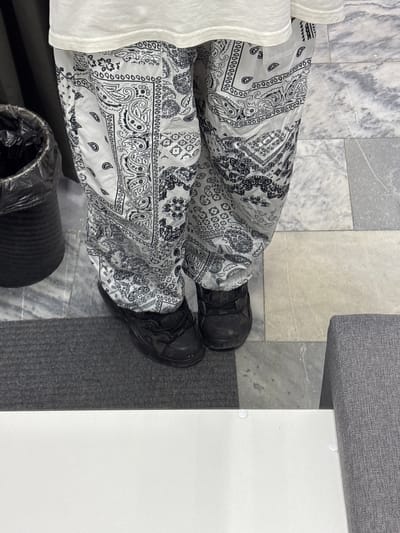 The image shows a close-up view of a person's legs wearing black patterned pants and black sneakers, standing on a tiled floor next to a dark gray rug. A black trash can is visible to the left of the person. The environment suggests an indoor setting, possibly a public space or a home. There is no indication of the city of Kuznetsk, Russia, or any other specific location.