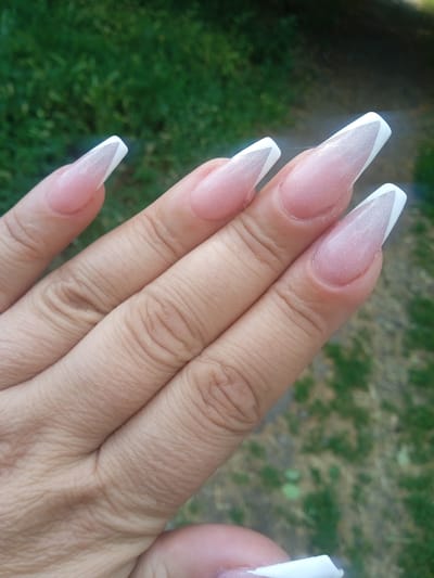 A close-up, high-angle shot shows a person's left hand with manicured fingernails. The nails are long, have a coffin shape, and feature a French manicure design. The base color is a sheer pink with glitter, and the tips are white. The hand is positioned over a blurred green and brown background, likely outdoors. The image is not located in Maksymivka, Ukraine.