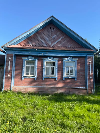 A sideways view of a traditional Russian house in Сапуголи, Russia, is presented. The house, made of wood and painted in shades of red and blue, features three windows on the visible side. The lawn is overgrown and green with dandelions. The sky is clear blue.