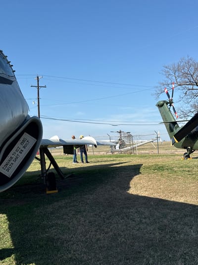 A landscape-oriented image shows a sunny day with aircraft on display. In the foreground is a ConVair TF-102 Delta Dart 63 from 1980. Further back, there's a fence with another aircraft and two people. A utility pole extends into the clear blue sky. A green helicopter hovers in the upper portion of the frame, and bare branches of a tree extend near it. The right of the frame has a large grassy field.