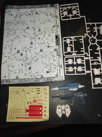 The scene is located in Mornington, Australia, displaying model kit components arranged on a dark, textured surface. A folded instruction manual lies on the left, open to pages illustrating assembly diagrams for various units including "Head & Body Unit," "Arm & Leg Unit," "Wing Unit," and "Weapons." To the right of the manual, multiple off-white plastic sprues hold numerous un-assembled model parts, with some individual pieces detached or partially detached from their frames. Small, dark, circular components, consistent with polycaps or screws, are scattered among these sprues. In the lower-left quadrant, a yellowish sheet of waterslide decals or stickers features a variety of small red, black, and white markings, including numerical identifiers such as "210" and decorative symbols. Below the sprues and to the right of the decal sheet, three distinct model components are present: a blue-grey missile-like weapon, a dark grey rifle, and a light beige/off-white shield or armor piece with dark accents. No individuals are visible in the image.