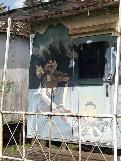 A weathered structure featuring a mural is visible. The mural portrays a man wearing a cowboy hat, seated and holding a cup. Beside him, there appears to be a bundle of wood or branches. To the man's right is a door with a covered window and a painted image of a lantern below. The entire structure seems old and in need of repair. A rusted, white metal railing is in the foreground, partially obscuring the view. Vegetation grows beneath the structure.