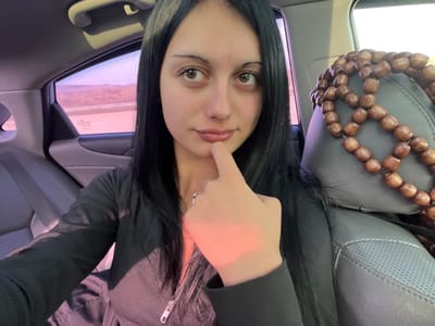 A young woman with long dark hair and light brown eyes is depicted in a close-up, chest-up view, seated in the rear passenger compartment of a vehicle. Her right index finger is positioned against her lower lip/chin area. She is wearing a black, long-sleeved, ribbed top with a visible zipper pull on the front and a thin silver-colored necklace with a small pendant. A ring is visible on her right ring finger.
The car's interior features dark grey upholstered seats, possibly leather or synthetic, with perforated sections visible on the front passenger seat headrest. This headrest is fitted with a brown wooden beaded seat cover. The car's interior trim is also dark grey. Through the left rear window, an exterior landscape is visible, showing a road or highway and distant, relatively flat, open terrain under a bright sky. The window glass has a strong purple/pink tint, affecting the color of the visible exterior. Strong, warm-toned light enters the vehicle from the right side, illuminating the subject's right side and hand.
The specific location within the city of Surami, Georgia, is not visually discernible from the details presented in the image's background.