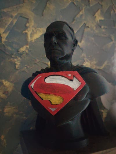 A dark, possibly black, bust of a man wearing a Superman emblem is displayed against a textured background. The bust is dark, with visible facial features like a nose and eyes, and appears to be made of a rigid material. The Superman emblem on the chest is a red shield with a yellow "S" and a white outline. A dark cape is draped over the shoulders. The background is a mottled pattern of grey and yellow, suggesting a wall or textured surface. The lighting is dim, casting shadows and highlighting the texture of the bust and background. The scene does not provide any specific clues to its location within the city of Сухарево, Russia.