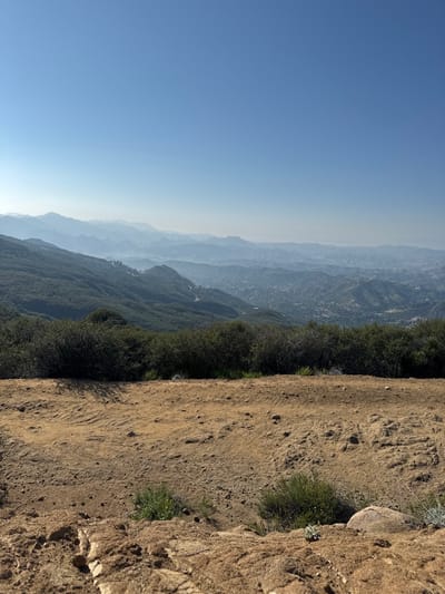 The image displays a panoramic view of a vast, arid mountainous landscape in the Unincorporated Santa Monica Mountains, United States of America, under a clear, pale blue sky.
In the immediate foreground, a broad expanse of reddish-brown, dry earth dominates the lower third of the frame. This ground is uneven, marked by subtle undulations, small rocks, and sparse patches of low-lying green and silver-hued vegetation.
Beyond the foreground, a dense, dark green band of bushy, chaparral-like vegetation forms a horizontal line, separating the immediate viewpoint from the expansive scenery beyond. This vegetation shows variations in density and height.
The middle ground and background reveal numerous layers of undulating hills and mountains, receding into the distance. These landforms are covered in a mix of green vegetation and exposed earth. As the mountains recede, they progressively lighten in color, appearing in shades of blue-gray due to atmospheric haze. Within the valleys and on the slopes of these distant hills, signs of human development are visible, including roads and clusters of buildings indicative of urban or suburban sprawl.
The sky, occupying the upper half of the image, is a bright, clear blue, transitioning slightly to a lighter hue near the horizon. A faint, white contrail is discernible in the upper-right quadrant of the sky. No people or animals are present in the scene. The overall environment suggests a dry, temperate climate with significant elevation changes and distant urbanization encroaching on natural terrain.
