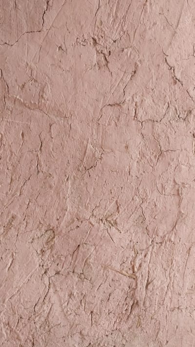 This is a close-up, full-frame, vertical shot of a cracked, textured, pinkish-brown mud wall. The surface is uneven with visible indentations and ridges, suggesting it is an unplastered or roughly finished building material. Fine, dark cracks web across the surface, varying in length and width, indicating dryness and age. Some lighter, straw-like fibers are embedded in the mud in various areas. The lighting is even, with no strong shadows, providing a clear view of the wall's texture. The image does not contain any people or discernible objects other than the wall itself. It is impossible to determine the specific location in Yangiobod, Tajikistan from this image alone, as it focuses solely on the surface detail of the wall.