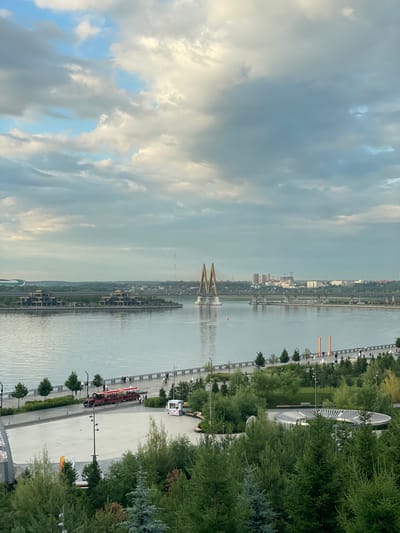The image captures a scenic view of the Kazanka River in the city of Kazan, Russia, looking towards the Millennium Bridge. The sky is filled with dramatic clouds, suggesting either early morning or late afternoon. On the river's left bank, there are modern buildings and lush greenery. A pedestrian walkway runs along the riverbank in the foreground, where several people are visible walking and one person appears to be operating a red tour train. Further across the river, the striking Millennium Bridge stands out with its distinctive golden-yellow pylons. Beyond the bridge, the cityscape of Kazan is visible, with various buildings and structures, including cranes indicating ongoing construction. The overall scene conveys a sense of urban development integrated with natural landscapes and recreational spaces.