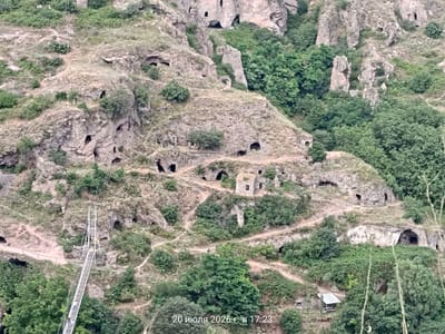 The image captures a panoramic view of the cave city of Khndzoresk, Armenia. In the foreground, a modern suspension bridge stretches across a ravine, with a few pedestrians visible on it. The bridge connects to a winding path that leads up the rocky hillside. The hillside is dotted with numerous natural caves and man-made structures carved into the rock face, suggesting a historical settlement. Some of these structures appear to be dwellings or chapels, with visible entrances and windows. The area is overgrown with lush green vegetation, including trees and shrubs, interspersed with dry grass and rocks. The overall impression is one of a rugged, historical landscape that has been adapted for human habitation over centuries. The date and time stamp "20 июля 2026 г. в 17:23" is visible in the bottom right corner.