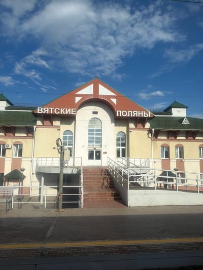 This is a daylight exterior, eye-level shot of the Vyatskiye Polyany railway station building in Vyatskiye Polyany, Russia. The building is constructed of light-colored brick and has a red, gabled roof with green trim. The name of the city, "ВЯТСКИЕ ПОЛЯНЫ," is displayed in large white letters on the front of the building. The building has multiple windows, including several large arched windows, and a central entrance with double doors. There are stairs and a ramp leading up to the entrance, with white railings on either side. The area in front of the building is paved with bricks and features a yellow safety line along the edge of the platform. A street lamp is visible to the left of the entrance, and a bench is also present on the platform. The sky above is blue with scattered white clouds. There are no people visible in the image.