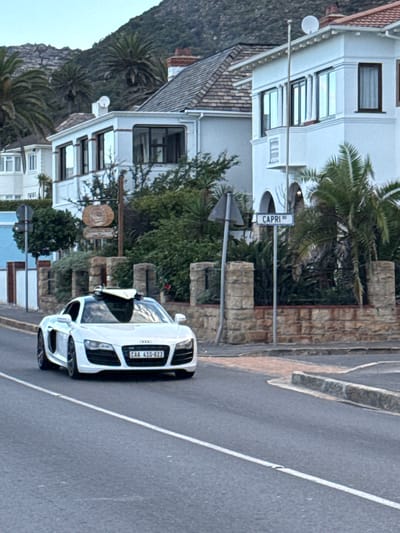 A white Audi R8 is parked on the roadside of Capri Road in Muizenberg, South Africa. The street is paved and has a white dividing line. Adjacent to the road is a stone wall with a street sign indicating "Capri Rd". Behind the wall is lush vegetation and a multi-story white building with dark-framed windows. In the background to the left, a mountain range is visible.