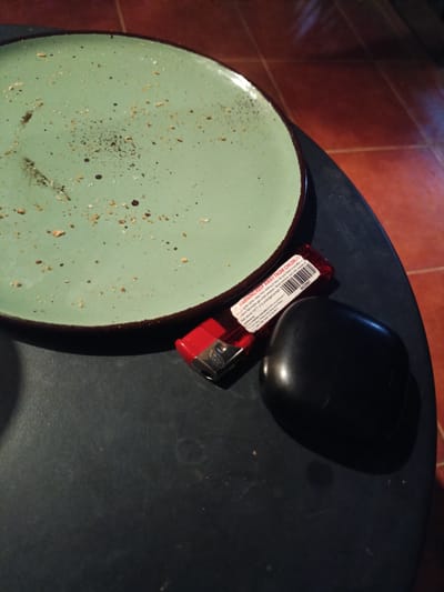 A close-up shot shows a light green plate with scattered crumbs and dark specks, likely from food. To the right of the plate, a red lighter is positioned, with a warning label visible. The label text includes "WARNING:KEEP AWAY FROM CHILDREN," information about flammable gas, and temperature restrictions. Beside the lighter is a smooth, black, oval-shaped object, possibly a stone or a piece of technology. The background is a dark, textured surface, possibly a table, and beyond that, a reddish-brown tiled floor is visible. The lighting suggests it might be daytime, with sunlight casting shadows and highlights on the objects. The overall scene appears to be a still life arrangement of everyday items, possibly on a table in a home or outdoor setting in Ban Patan, Thailand. There are no people visible in the image.
