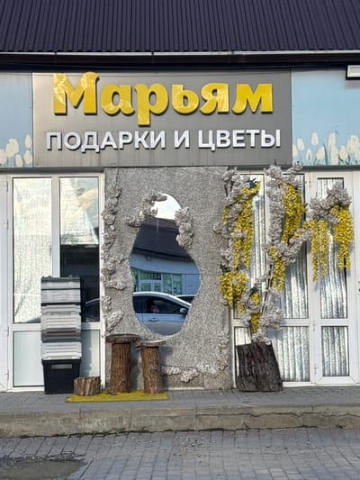 The image shows the exterior of a shop in Kurchaloy, Russia, with the name "Марьям" (Maryam) displayed in large yellow letters above the words "ПОДАРКИ И ЦВЕТЫ" (Gifts and Flowers) in smaller white letters. The storefront features a large, irregularly shaped cutout in a textured grey facade, through which a reflection of a street with a car and buildings is visible. Artificial yellow and white flowers hang from the facade around the cutout and extend to the right. To the left of the cutout, stacked grey crates and a black bin are placed near a glass door. In front of the facade, on the sidewalk, are three tree stumps of varying heights, one with a flat top, placed on a small patch of artificial yellow grass. A larger piece of wood is positioned to the right of the facade, near another glass door. The ground is paved with bricks, and a small puddle of water is visible in the foreground. The overall scene depicts a commercial establishment with decorative elements.