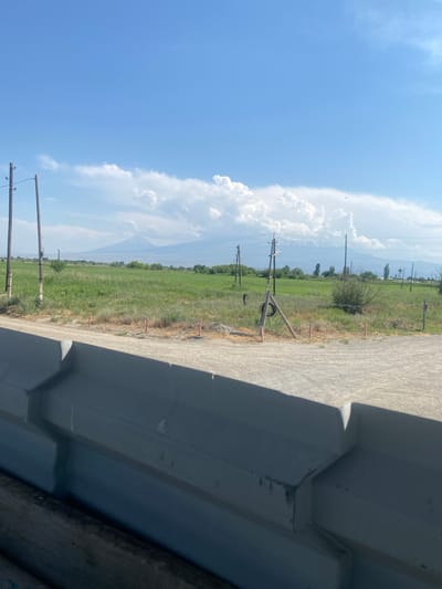 This media file depicts a rural landscape under a bright, sunny sky. The image is oriented vertically, showing a wide expanse of green fields and a dirt road. Several utility poles with wires traverse the scene, suggesting the presence of infrastructure in a relatively undeveloped area. A large, grey, angular structure dominates the right side of the frame, possibly a modern building or a section of a bridge. The sky is a vibrant blue, with large, fluffy white clouds accumulating on the left side, indicating fair weather with potential for cumulus cloud development. The sun appears to be high in the sky, suggesting it is midday. The overall mood is serene and somewhat open, with a sense of quiet rural life. There is no visible text in the image. The location context provided is Ranchpar, Armenia.