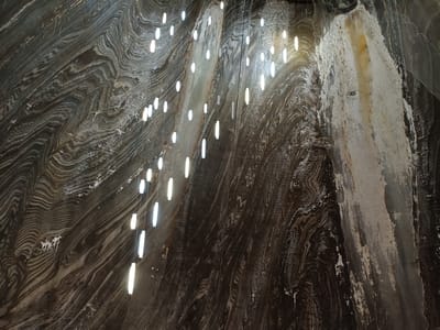 The image shows a cavernous space with walls made of striated salt. The striations are in varying shades of dark brown, grey, and white, creating a swirling, organic pattern. Numerous vertical, rod-shaped lights are suspended from the ceiling by thin wires, illuminating the salt formations and casting a soft glow. Some of the lights appear clustered together, while others are more spread out, creating a dynamic visual effect. The salt walls show textures of both smooth, polished surfaces and rough, crystalline patches. There are no people visible in the image. The overall impression is one of a natural underground environment enhanced by artificial lighting, likely a tourist attraction. The lighting suggests it could be day or night, as it is an underground location. The location cues point to a salt mine, specifically Turda Salt Mine in Romania, known for its large chambers and unique geological formations.