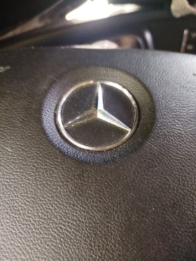 The image shows a close-up view of a Mercedes-Benz logo on a black leather steering wheel. The logo is circular with a three-pointed star in the center, and it appears to be made of chrome. The steering wheel is textured, with a visible grain pattern on the leather. The lighting suggests the photo was taken indoors, possibly in a car parked in a dimly lit area. There are no people visible in the image, and the background is out of focus, showing some blurred shapes that might be parts of a car's interior. The exact location in Braintree, United Kingdom, is not discernible from the image itself.