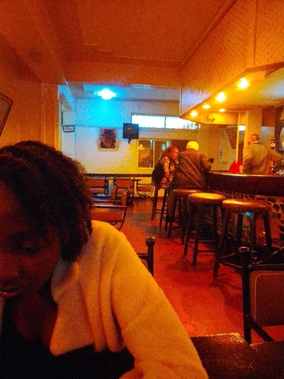 The image captures an interior scene, likely a bar, pub, or a dimly lit restaurant, characterized by warm, reddish-orange artificial lighting that bathes most of the space. The overall atmosphere appears casual and social, set in the evening or night.
In the immediate foreground, on the left side of the frame, a person with dark, curly hair is partially visible, looking downwards or away from the camera. They are wearing a light-colored, possibly off-white or pale yellow, long-sleeved top or cardigan over a darker garment. A dark, reflective tabletop is visible in the very bottom right corner, suggesting the person is seated.
Towards the midground and right side of the image, a prominent bar counter with a dark top and decorative tiling on its side can be seen. Several dark wooden bar stools are placed along the counter. Three individuals are gathered around the bar area:
* One person, wearing a dark jacket, is seated on a stool with their back partially to the camera, facing the bar. They appear to have light-colored hair or headwear.
* Next to them, another person with dark skin is standing or seated on a stool, turned slightly towards the left, seemingly interacting with the individual beside them or the bartender (who is not clearly visible). They are wearing a patterned top or jacket.
* Further down the bar to the right, a third person is vaguely visible, also seated with their back to the camera.
A small red object, possibly a bag or cloth, rests on the bar counter. Above the bar, a fixture with multiple warm, yellowish lights illuminates the area.
Beyond the bar area, in the background, a seating arrangement of tables and chairs is visible. The chairs feature dark frames and reddish-orange upholstered seats. The floor throughout the establishment appears to be a solid reddish-brown or terracotta color. On the back wall, a framed picture or artwork is hanging, with a black speaker mounted below it. A small, rectangular white object, possibly an electrical fixture or a sign, is also present on the wall to the left of the artwork.
A striking feature in the background is a bright, luminous blue light source located high up, contrasting sharply with the dominant warm tones of the rest of the room. This could be a decorative light or light spilling from an adjacent space. A window or doorway is also visible in the far background, letting in a subtle amount of light from outside, though it's largely obscured. The ceiling has a textured surface, possibly wood paneling, reflecting the warm glow of the lights.
The image has a noticeable grainy texture, characteristic of photos taken in low-light conditions, possibly with a higher ISO setting. No discernible text is visible in the frame.