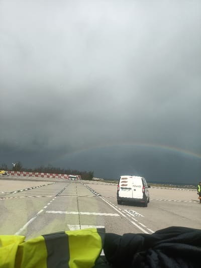 A rainbow arches over an airport runway under a stormy, overcast sky. A white van with “NEWPCS” on its side drives on the runway. The runway has white lines and markings. A bus is parked on the left side. A person in a yellow safety vest stands on the right side. The foreground shows parts of a yellow safety vest.