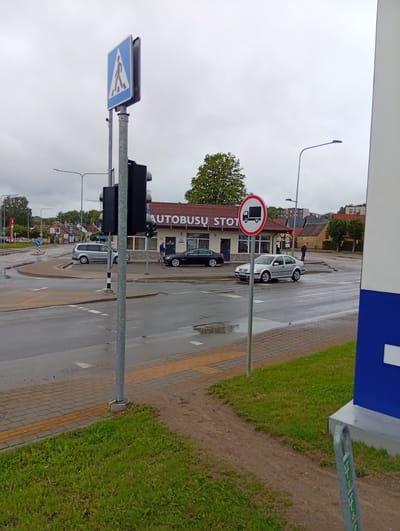The image depicts a street scene in Kuršėnai, Lithuania, likely near a bus station as indicated by the sign "AUTOBUSU STOT." A cloudy sky suggests overcast weather. In the foreground, a paved walkway with tactile paving leads to a grassy area. A traffic light pole with a pedestrian crossing sign is prominent on the left. Several cars are visible: a silver minivan in the left lane of a roundabout, a black sedan in the right lane of the roundabout, and a silver sedan in the right lane of the road in the middle ground. A circular traffic sign with a red border and a black truck symbol on a white background is affixed to a pole to the right of the silver sedan, indicating a prohibition for trucks. The bus station building has a reddish-brown tiled roof and visible windows. One person, a male wearing a blue top, is standing near the entrance of the bus station. Another person, a male wearing dark clothing, stands further back on the sidewalk. The road surface appears wet, suggesting recent rain.
