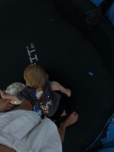 A child with blond hair is lying on a black trampoline. The child is wearing a dark t-shirt with cartoon characters on the back. Next to the child are two soccer balls. A person's hand and foot are visible in the lower left and right of the frame, respectively. The trampoline has blue padding on the sides, some of which appears torn. The scene is outdoors, and the lighting suggests it is daytime.