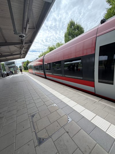 The image shows a train platform at Gronau (Westfalen) station in Gronau, Germany. A red and silver train is stopped at the platform. The platform is paved with grey rectangular tiles and has a tactile paving strip along the edge. A covered walkway with a metal roof extends over part of the platform. A security camera is visible mounted on the underside of the walkway roof. In the background, there are trees and buildings. A few people are visible on the platform, including one person running and another person with a stroller.