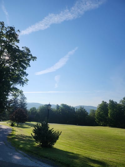 A bright, sunny day in the Town of Hoosick, United States of America, is depicted with a clear blue sky adorned with wispy white contrails. The foreground features a paved pathway curving to the left, casting dappled shadows on the adjacent vibrant green lawn. A dark green, bushy shrub sits prominently on the edge of the lawn near the path, with a tall, dark lamppost standing behind it. Beyond the lawn, a line of dense green trees stretches across the middle ground, partially obscuring a distant building with a dark roof. In the far background, a range of blue-hued mountains forms the horizon, softened by the atmospheric haze. A large, leafy tree with sunlight filtering through its branches dominates the upper left quadrant of the image.