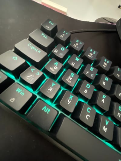 The image shows a close-up of a black keyboard with green backlighting. The perspective is from above and to the side, focusing on the keys. The keyboard appears to be a gaming keyboard, with some keys featuring symbols that are common in gaming or international layouts, such as Cyrillic characters and symbols like "Mac" and "Win". Some of the keys are clearly labeled with their function or character: "Esc", "Tab", "CapsLk", "Win", and "Alt". The overall scene is a still life of an electronic device, with no people or specific events depicted. The lighting suggests that the image was taken indoors, possibly in a room with low ambient light to highlight the keyboard's backlighting. There are no obvious clues to indicate the time of day, weather, or a specific location beyond the assumption that it is an indoor setting. The texture of the keyboard and the surrounding surface is visible, with some dust particles present on the black surface. The crops provided focus on different sections of the keyboard, further detailing the inscribed characters on the keys.
