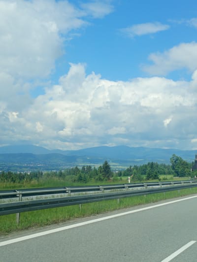 The image captures a daytime scene along a roadside in Rdzawka, Poland, looking towards rolling hills and mountains under a partly cloudy sky. The foreground features a grey asphalt road with a white lane marking, bordered by a metal guardrail. Beyond the guardrail, a strip of green grass and foliage lines the embankment, with various trees and a dense green forest extending towards the middle ground. A small cluster of buildings, possibly a village or town, is visible nestled in the valley between the trees and the distant mountains. The background is dominated by layered mountain ranges that fade into a hazy blue. The sky is a bright blue with scattered white cumulus clouds. There are no people or animals visible in the frame. The overall impression is a scenic view of a rural landscape observed from a roadway.