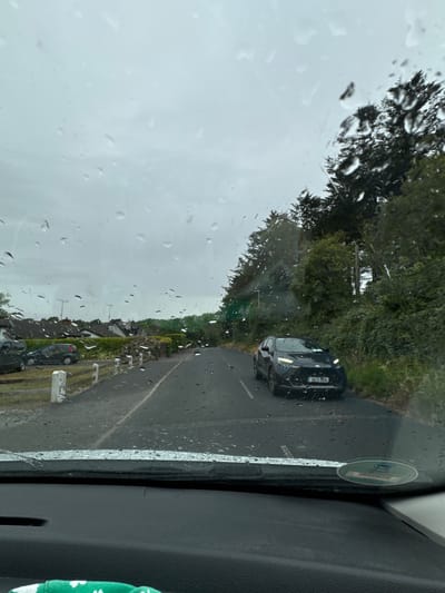 The image is taken from inside a car looking through a rain-covered windshield on a cloudy day. The scene depicts a road in Courtown, Ireland. On the left side of the road, several parked cars are visible, along with a white picket fence and houses in the background. The road itself is wet, with visible puddles and streaks from the rain. A dark-colored SUV is driving on the right side of the road, facing the camera. Trees and foliage line the right side of the road, forming a dense border. The overall environment suggests overcast weather conditions.