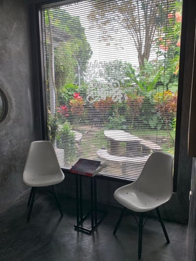 The image depicts an interior seating area facing an outdoor garden, likely located at The Cottage in Kota Batu, Indonesia. The interior space features dark grey, smooth, possibly polished concrete walls and floor. A large, rectangular window with a black frame and horizontal white Venetian blinds dominates the central view. Two white, modern, molded plastic chairs with splayed black legs are positioned in the foreground, facing each other. Between the chairs, directly in front of the window, is a small, black, rectangular metal C-table. On this table, a red-covered board game box, possibly "Monopoly," rests atop a white book. A circular, black-framed mirror is visible on the left concrete wall.
Through the window's blinds, a vibrant, well-maintained garden is visible. Key features in the garden include lush green foliage, bright red and orange-brown-leaved plants, and taller trees in the background against a light sky. White, raised letters spelling "THE COTTAGE" are arranged as a landscape feature within the garden. A light-colored, curved concrete or stone bench and an associated table are situated in the right-center of the garden, accessible via a visible path. Pink or orange hanging floral elements are visible on the far right. No individuals are present in the scene, and no actions or interactions are depicted.