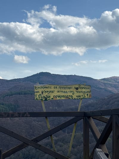 Here's a description of the image:
The image shows a partially visible sign attached to a wooden structure, possibly a railing or lookout point. The sign is in Bulgarian and reads: "Емоциите се превръщат в спомени, но боклукът остава сред природата! Пази природата чиста!" meaning "Emotions turn into memories, but trash remains in nature! Keep nature clean!"
In the background, there is a mountain range covered in dense vegetation, likely trees. To the left of the image, there is a blue sky with prominent white clouds.
Given the language on the sign, and that I know this is located in Delchevo, Bulgaria, I can infer that the location is likely in a natural, mountainous area in or around Delchevo.