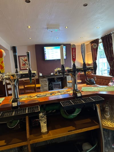 Inside a pub, beer taps stand on the bar, displaying brands like "Fosters" and "Amstel." A television screen displays the drink menu behind the taps. The bar's wooden structure houses beer lines and equipment. Soft lighting illuminates the room. The atmosphere appears cozy and inviting.