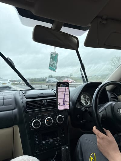 The image depicts the interior of a car, specifically the driver's side. A person is visible holding the steering wheel. A phone is mounted on the dashboard displaying a navigation app, showing directions to Barrow-in-Kendal and Kirkby Lonsdale. The time on the phone is 15:33. The car is in traffic, based on the view through the windshield. A car air freshener is hanging from the rearview mirror, and a Blaupunkt radio is visible. The weather appears overcast.